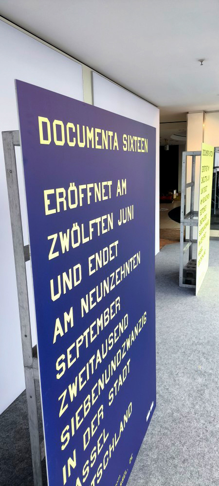 Großformatige blaue Hartschaumplatte mit gelber Typografie zur Ankündigung der documenta 16